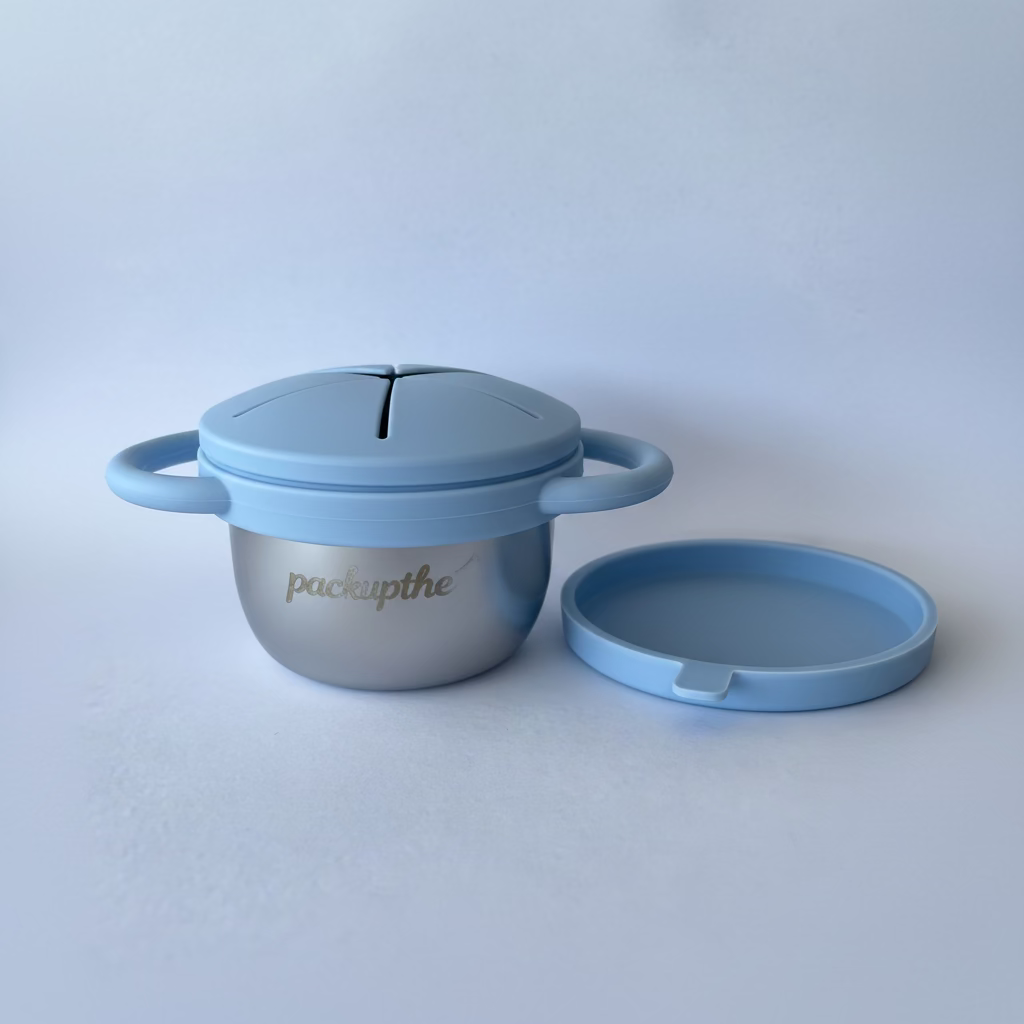 A light blue silicone baby feeding bowl with a matching lid, featuring a silver stainless steel inner bowl and the brand name 'packupthe' printed on the front. The lid is propped up behind the bowl on a light blue background.
